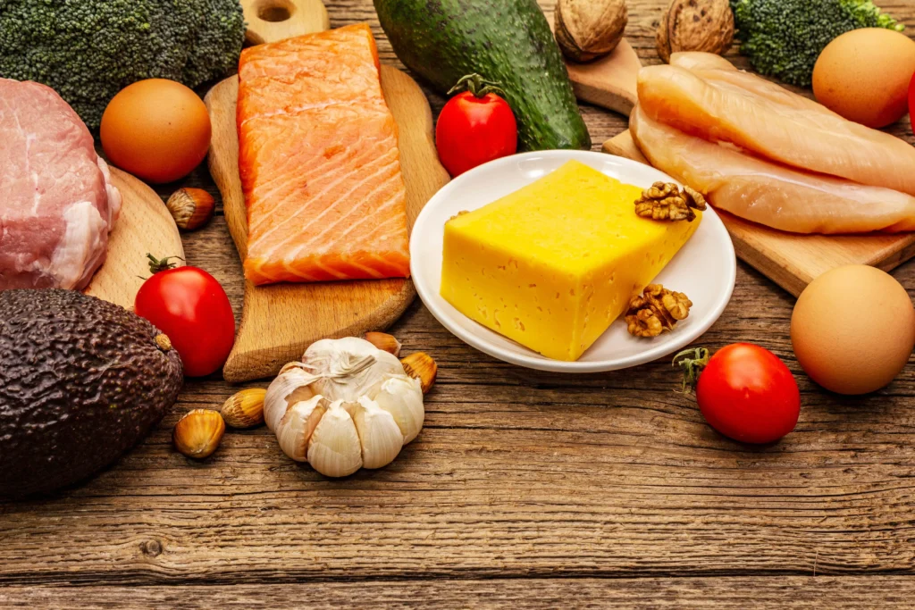 A variety of fresh food items arranged on a wooden surface, including salmon fillets, chicken breast, cheese, avocados, eggs, broccoli, tomatoes, and garlic.