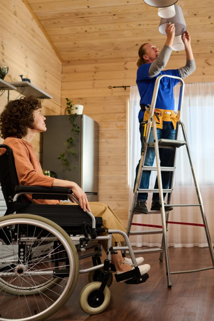A woman sitting in a wheelchair indoors watches a man standing on a stepladder while he changes a light bulb overhead.