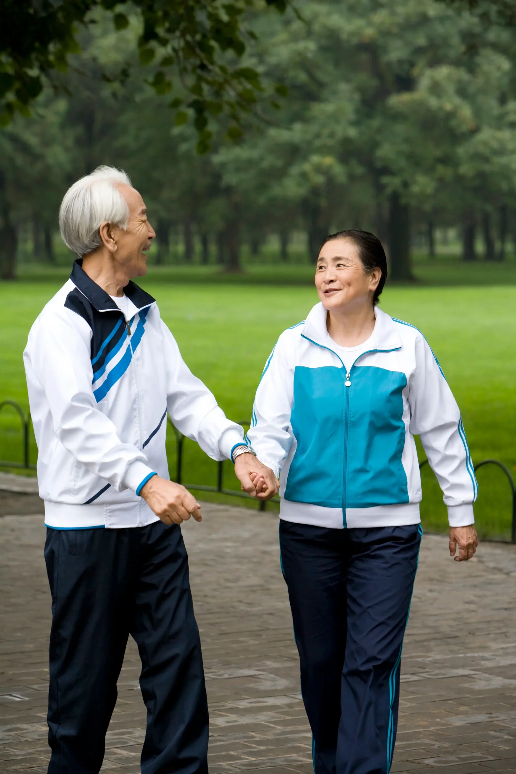 An older couple holding hands and walking together in matching tracksuits through a green park.