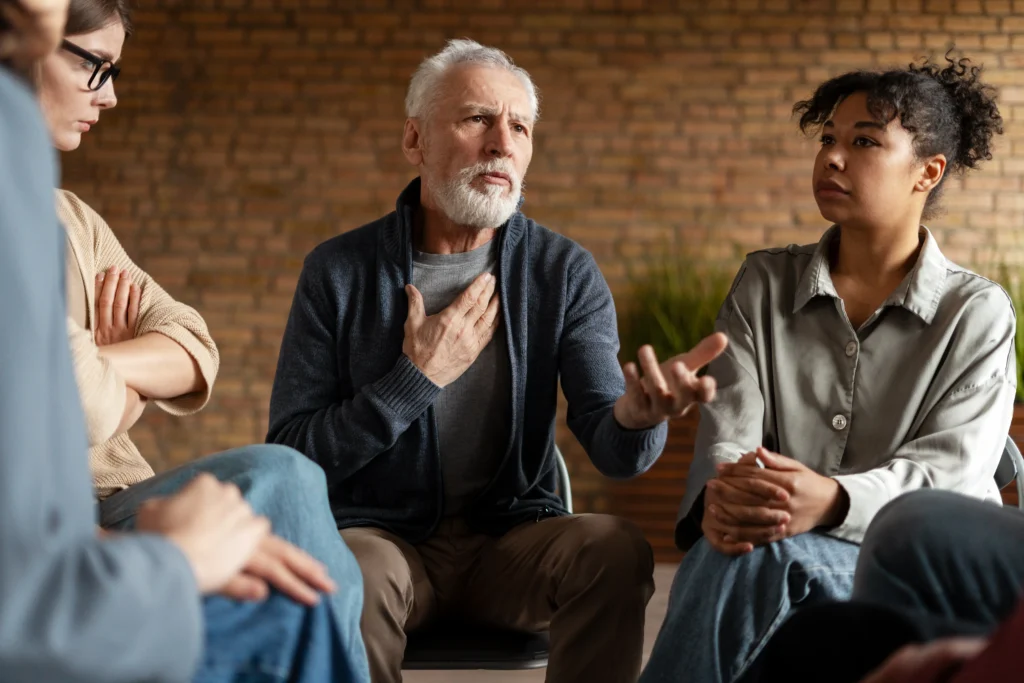 A senior man with a grey beard speaks attentively while gesturing with one hand to a group sitting in a circle during a support or discussion session.