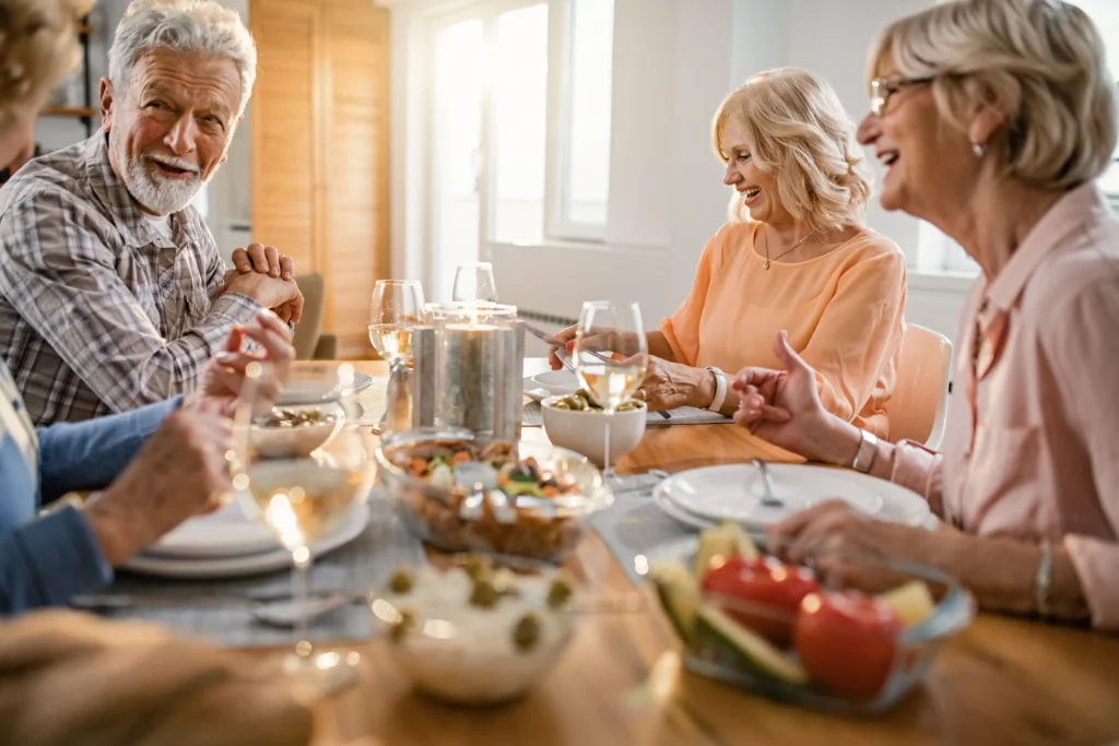 A group of cheerful senior friends sitting around a bright dining table, chatting and enjoying a meal together with wine.