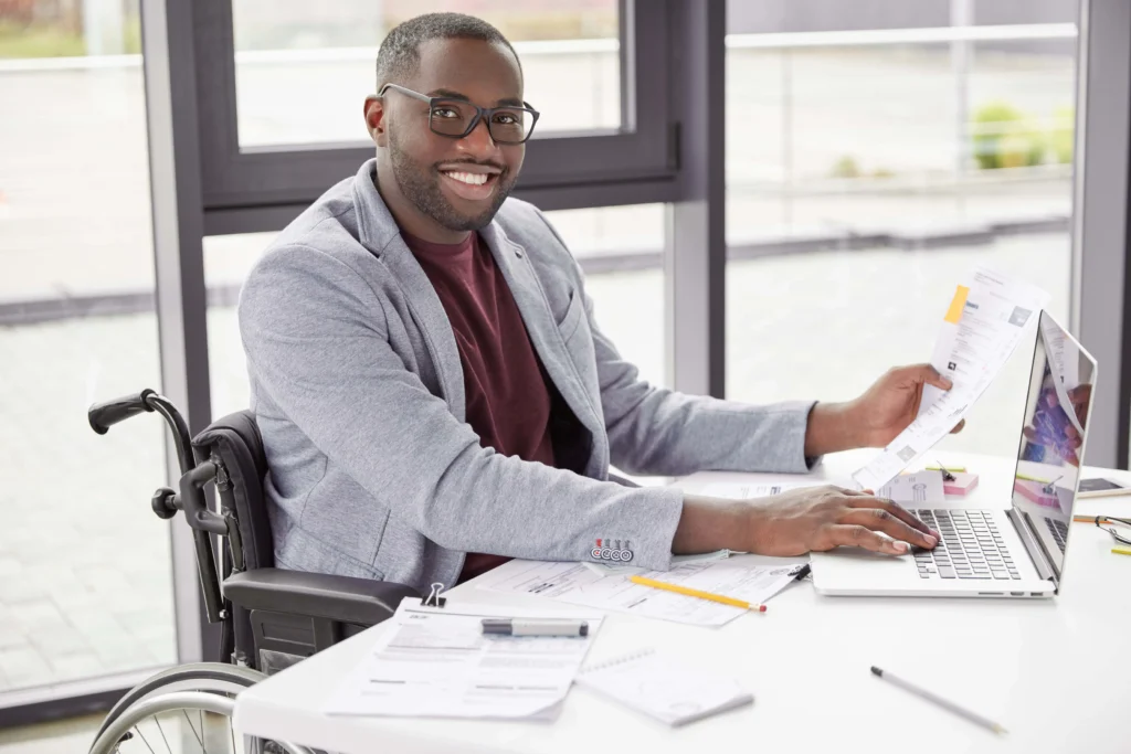 A smiling Black man wearing glasses, a grey blazer, and a maroon t-shirt sits in a wheelchair at a bright white desk, using a laptop and holding paperwork with modern office windows in the background.