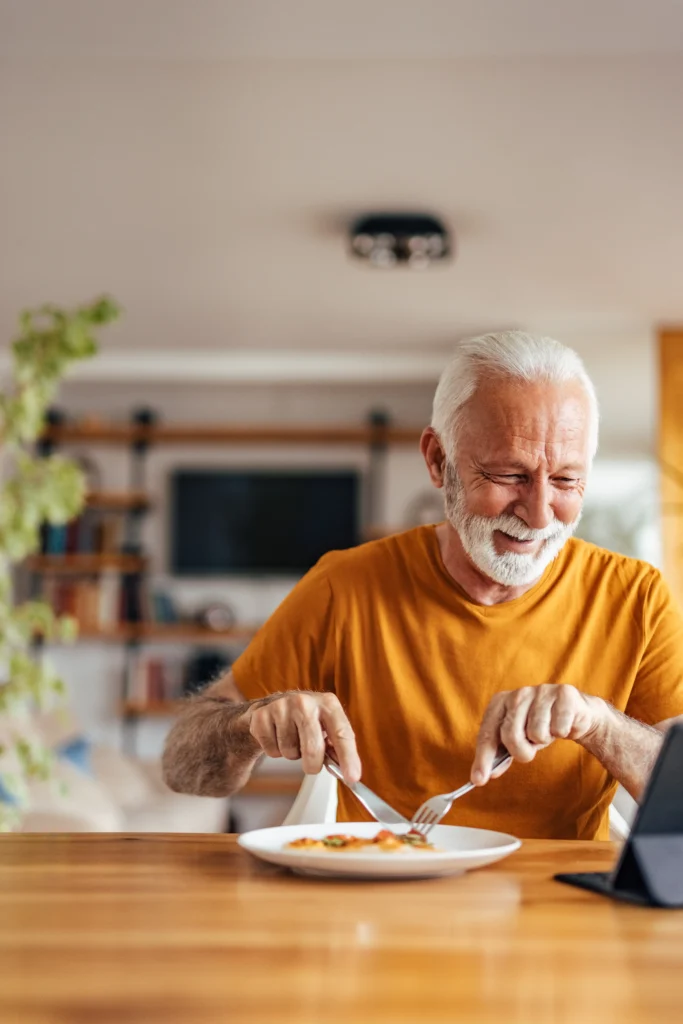 A happy senior man with a white beard smiling while cutting into a meal at a wooden table while watching a digital tablet.