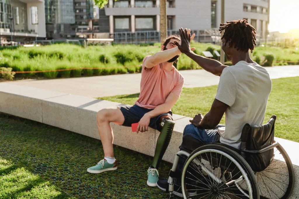 Two friends high-five outdoors in a sunlit park setting—one man seated on a low concrete wall wearing a pink t-shirt and showing a prosthetic leg, and the other man sitting in a wheelchair seen from behind with dreadlocks.