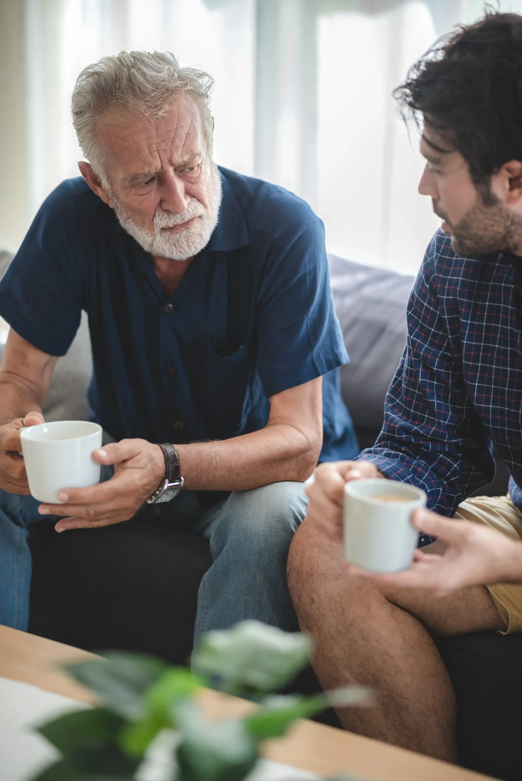 An older man and a younger man having a serious conversation while holding coffee mugs on a couch.