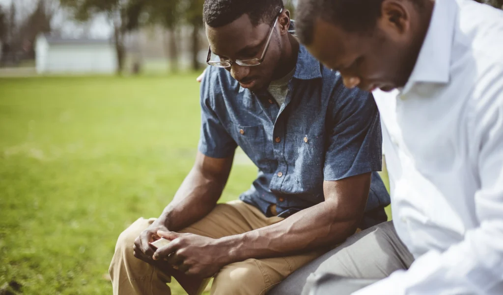 Two Black men sitting outdoors on a bench, looking down thoughtfully while one holds a small book or phone.