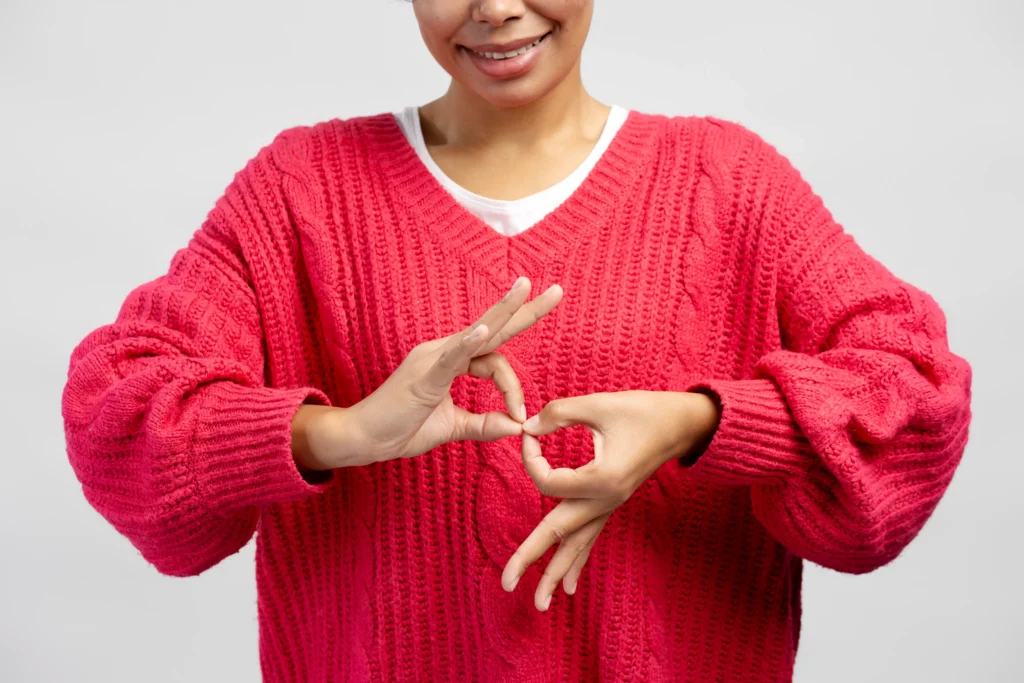 A close-up view of a woman wearing a bright pink knit sweater, using her hands to form sign language gestures against a plain white background.