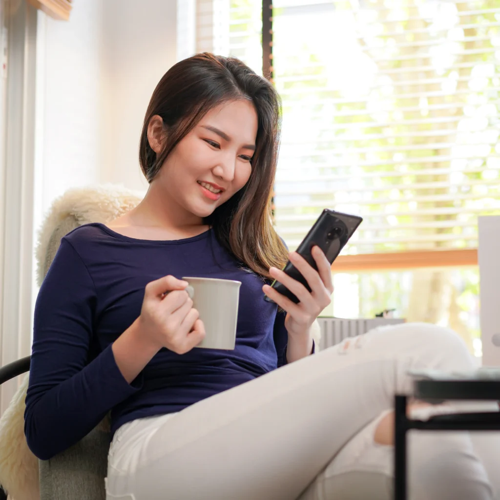 A young East Asian woman smiling while sitting in an armchair, holding a light grey mug in one hand and looking at her smartphone in the other near a brightly sunlit window.