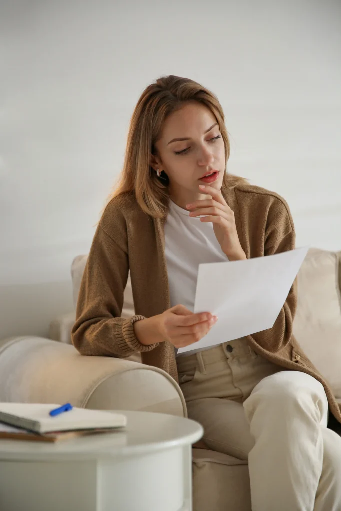 A woman sitting on a beige couch at home attentively reviews a white sheet of paper held in her hands.