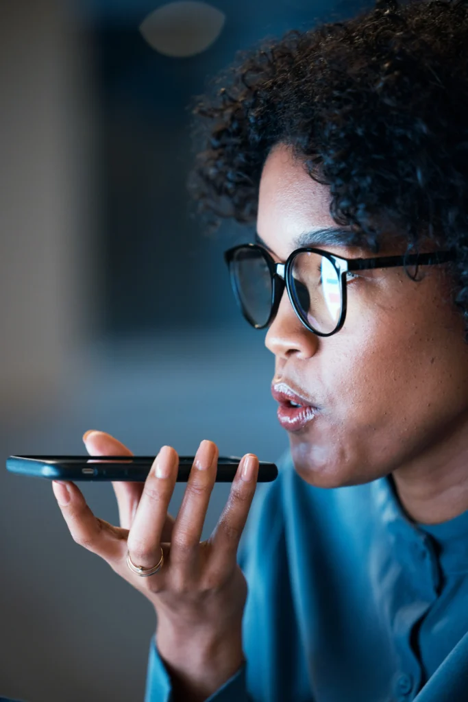 Close-up of a Black woman wearing glasses speaking directly into the microphone of a smartphone held horizontally near her mouth.