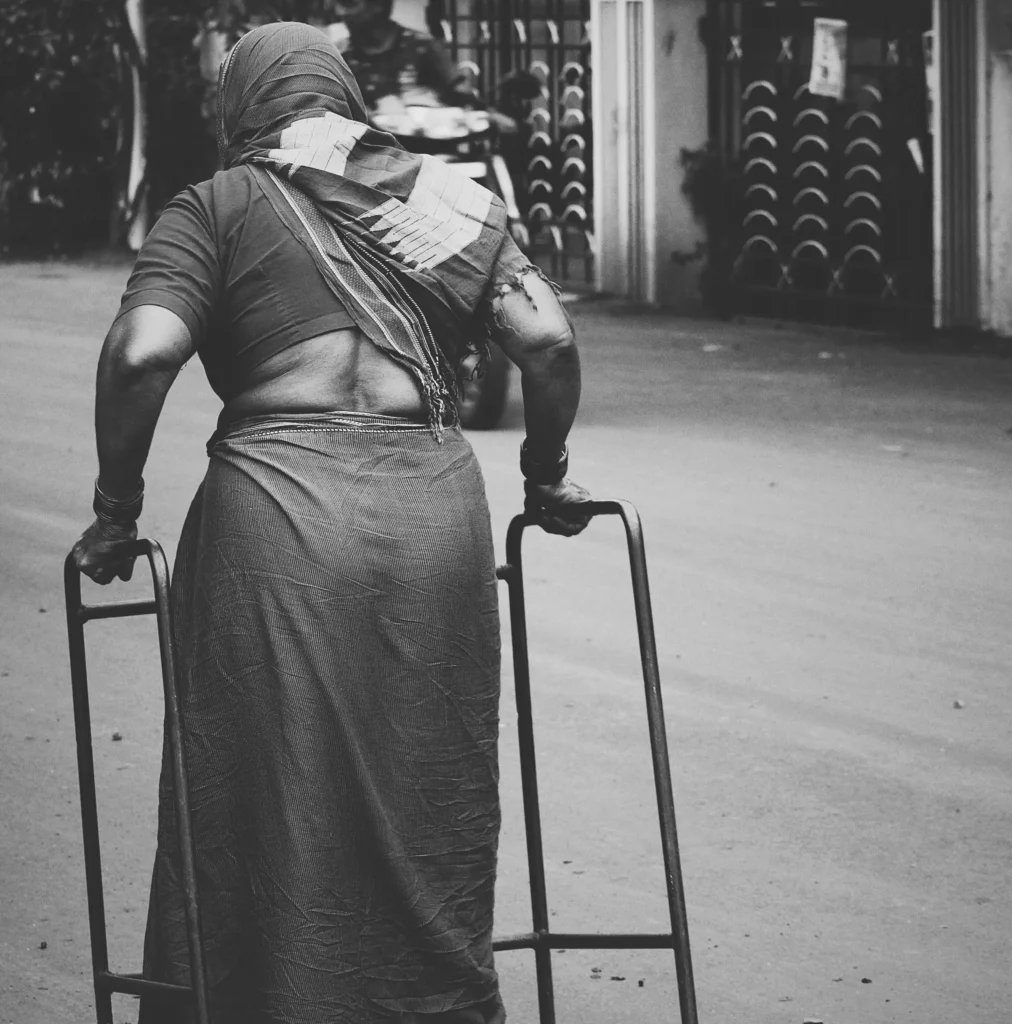 A black-and-white rear view of an elderly woman wearing a sari, using a walker frame to travel down a street.