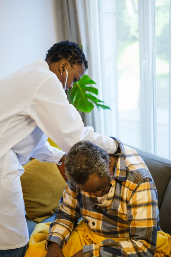A doctor in a white lab coat using a stethoscope to examine an older man sitting on a sofa.