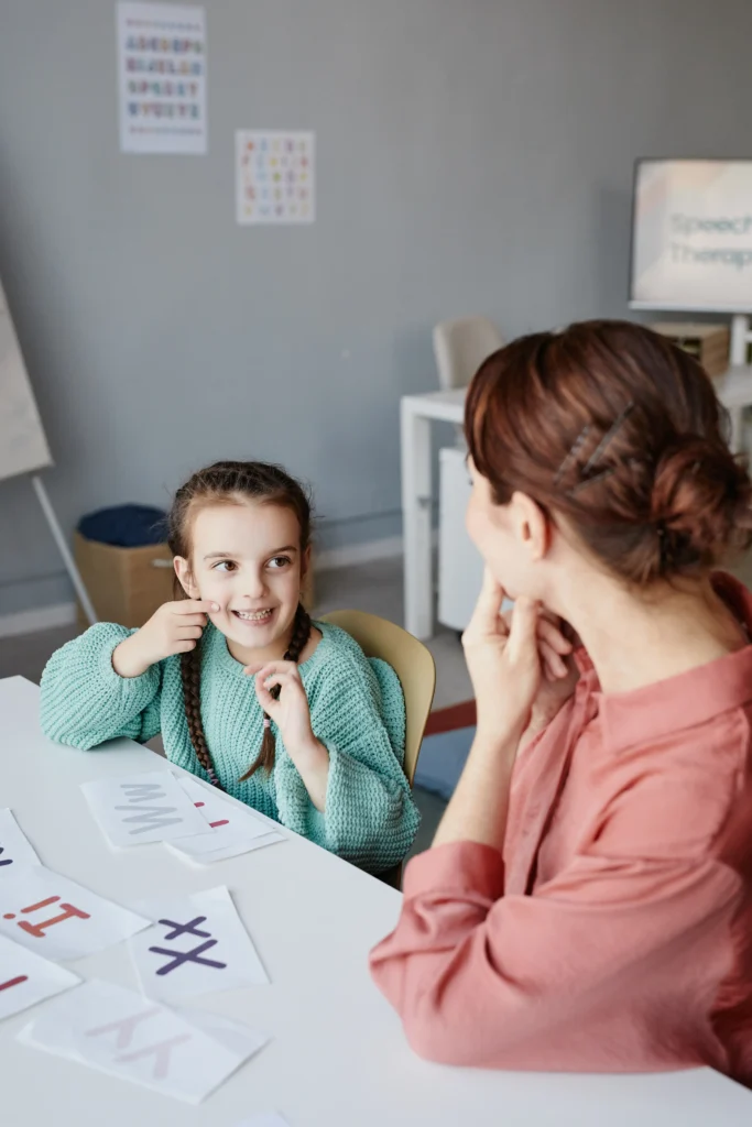 A young girl sitting at a table with letter flashcards, pointing to her cheek while mirroring a speech therapist or instructor seated across from her.