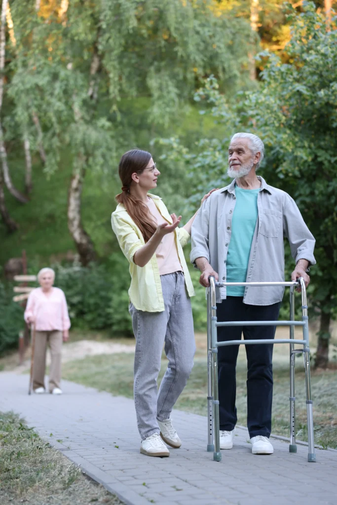 A young woman talking with a senior man using a walker on a paved outdoor path.
